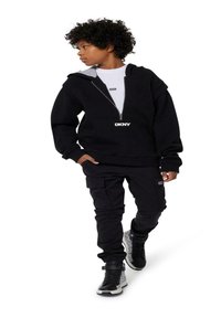 Felpa nera con zip frontale, foderata in grigio e con logo DKNY; abbinata a pantaloni cargo neri e scarpe alte con un design a fantasia.