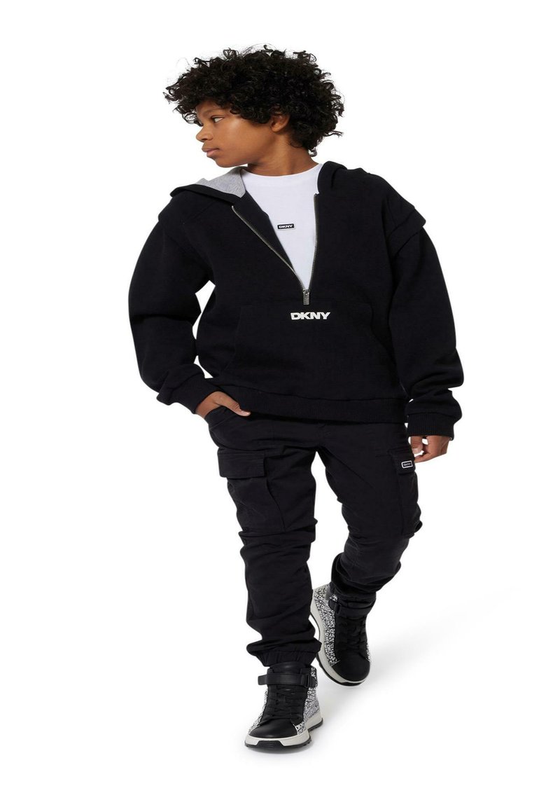 Felpa nera con zip frontale, foderata in grigio e con logo DKNY; abbinata a pantaloni cargo neri e scarpe alte con un design a fantasia.