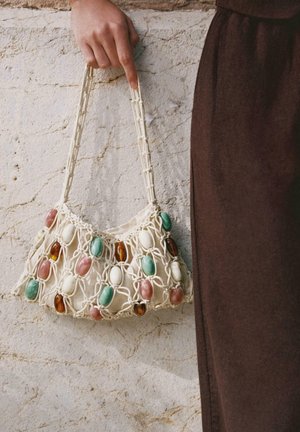 Mână ținând o geantă bej împletită cu mărgele ovale verzi, albe, roz și maro, în fața unui perete alb texturat și a unui articol vestimentar maro.
