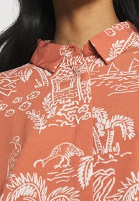 Luźna bluzka koralowa z kołnierzykiem zapinanym na guziki, zdobiona białymi wzorami kwiatów i motywami natury, w tym ptakami i liśćmi palmowymi, wykonana z teksturowanej tkaniny.