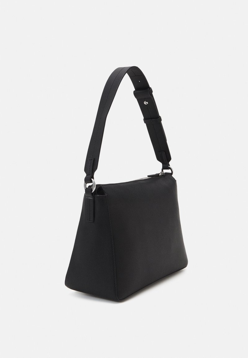 Sac à main noir en forme de trapèze avec bandoulière large réglable et fermeture éclair, présenté légèrement en angle sur un fond clair uni.