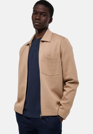 Jeune homme portant une veste beige zippée par-dessus une chemise bleu marine boutonnée, debout avec une main dans la poche devant un fond uni.