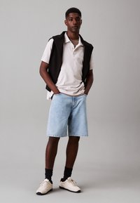 Polo shirt grigio chiaro, hoodie senza maniche nera, pantaloni shorts in denim blu e sneakers beige con suole scure. Design semplice, vestibilità casual, senza stampati.