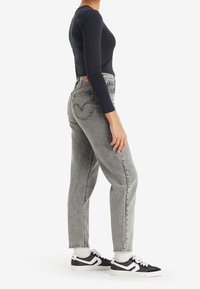 Una mujer se presenta de lado, vistiendo unos jeans grises de cintura alta, una camiseta negra de manga larga, calcetines blancos y zapatillas negras y blancas, todo ello sobre un fondo blanco.
