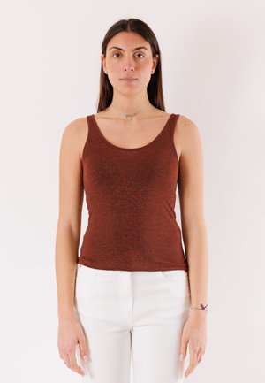 Giovane donna con lunghi capelli scuri, indossa un top senza maniche color ruggine e pantaloni bianchi, in piedi davanti a uno sfondo chiaro e uniforme.