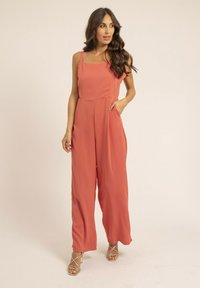 Combinaison corail avec décolleté carré, fines bretelles et design à jambes larges. Fabriquée en tissu léger, avec des poches latérales et une texture lisse.