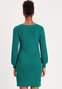 Robe tricotée couleur bleu sarcelle avec manches longues, texture côtelée et silhouette ajustée. Caractérisée par un large décolleté et des détails subtils aux poignets.