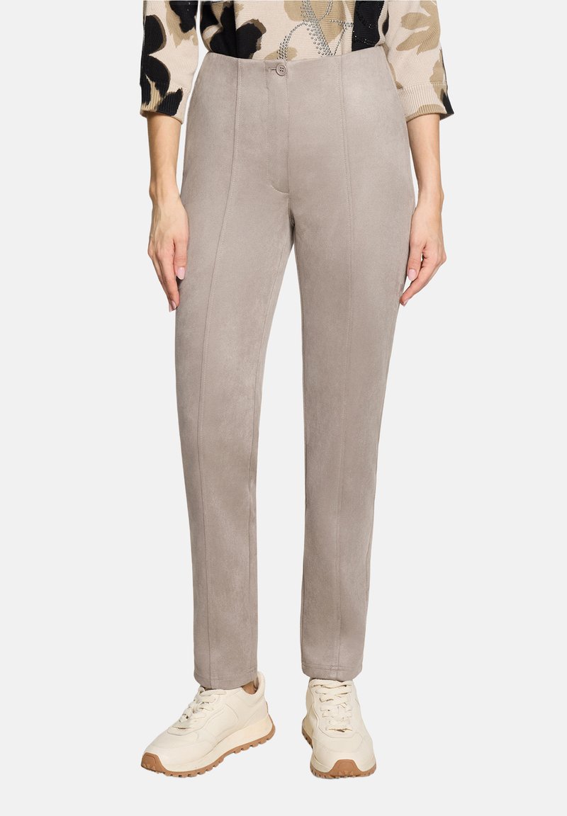 Femme portant un pantalon slim gris clair et des baskets beiges, mains relâchées sur les côtés, avec un haut à motif partiellement visible.