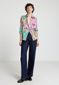 Colorful, patterned silk blouse in pink, green, and gold, paired with high-waisted dark denim pants and black shoes. Smooth texture, relaxed fit.
