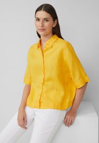 Chemise jaune en lin à manches courtes avec un col et une fermeture à boutons sur le devant. Le tissu a une apparence texturée. Portée avec un pantalon blanc.