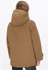 Bruine hoodiejack met een losse pasvorm, duurzame stof en zichtbare stiksels. Kenmerken zijn een rechte zoom en een enkele knoopmanchet.