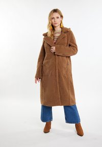 Brun, struktureret, lang frakke med hætte, store forlommer og en strømlinet silhuet, båret over blå opsmøgede jeans og brune ankelstøvler.