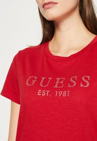 Raudona trumpų rankovių marškinėlių su minkšta medžiaga, ant krūtinės puošti sidabro spalvos karoliukais "GUESS" ir "EST. 1981". Apvalus iškirpimas.