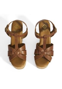 Sandales compensées en cuir marron avec un design de brides croisées complexe, une sangle de cheville réglable et une semelle en liège. Logo doré en relief visible.
