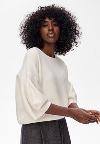 Weißer, übergroßer Pullover mit rundem Halsausschnitt und Puffärmeln, aus einem weichen Material, kombiniert mit dunklen, strukturierten Hosen.