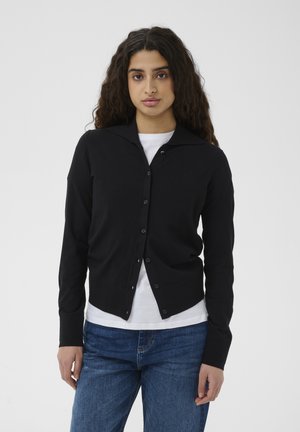 Hosszú, göndör hajú nő fekete, gombos kardigánban, fehér inggel és kék farmerrel, egyszínű, világos háttér előtt állva.