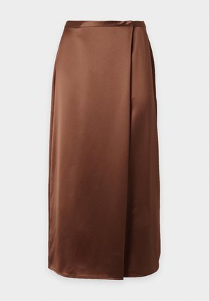 Femme debout portant un haut sans manches en satin marron et une jupe longue assortie avec des chaussures noires sur un fond clair uni.