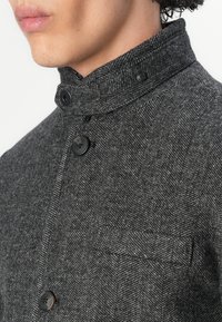 Veste gris à motif chevron avec un col haut, dotée de boutons noirs et d'une petite poche poitrine, fabriquée en tissu texturé.