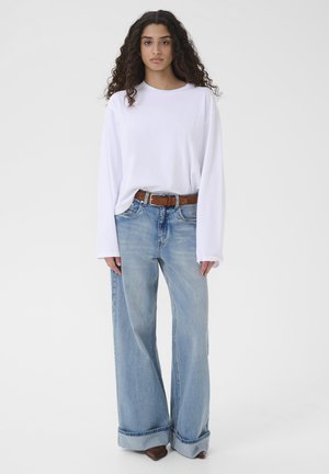 Žena s dlhými kučeravými vlasmi, oblečená v bielej blúzke s dlhým rukávom, hnedým opaskom, svetlomodrých širokých džínsach a hnedých špicatých topánkach.