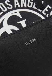 Czarna torebka ze skóry z fakturą, z srebrnym logo „GUESS”, z dużym białym nadrukiem graficznym na jednej stronie i zamkiem błyskawicznym.