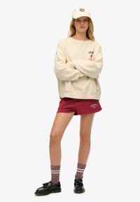 Joven de pie con los brazos cruzados, vistiendo un sudadera beige y una gorra, pantalones cortos burdeos, calcetines con estampado y mocasines negros con borlas.