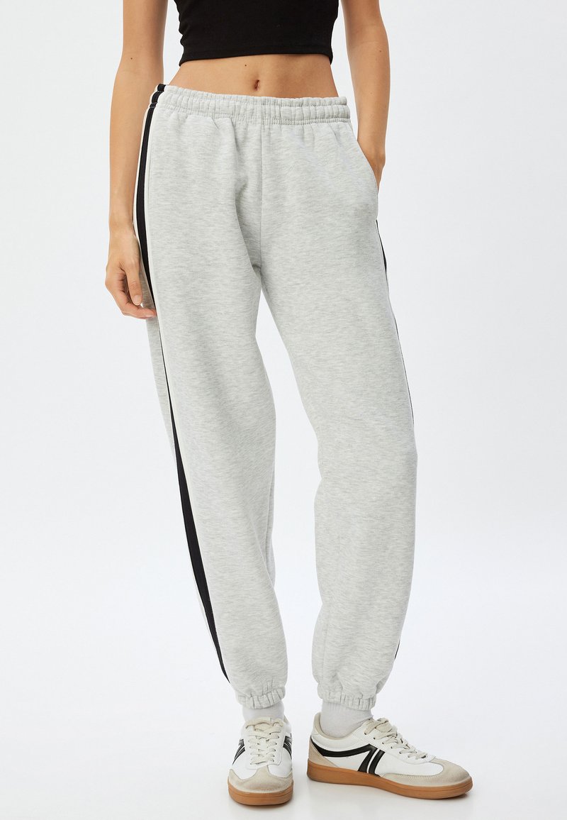 Grijze sweatpants met zwarte zijstrepen, elastische tailleband en taps toelopende boorden. Draagt witte en beige sneakers.