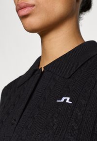 Polo negro de punto con un diseño de trenzado texturizado, que presenta un logo bordado en blanco en el pecho y un cuello clásico.