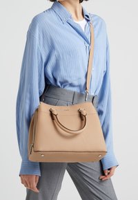 Beige strukturerad läderhandväska med dubbla topphandtag och avtagbar axelrem, med dragkedjestängning och metallnitar vid basen.