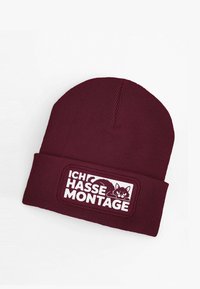 Burgundy knit beanie featuring a white patch with the words "ICH HASSE MONTAGE" and an illustration of a black cat. Soft texture, close-fitting design.