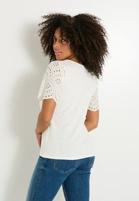 Camiseta blanca con mangas cortas de encaje de ojales y un escote redondo medio, combinada con jeans azules. Textura de tela suave.