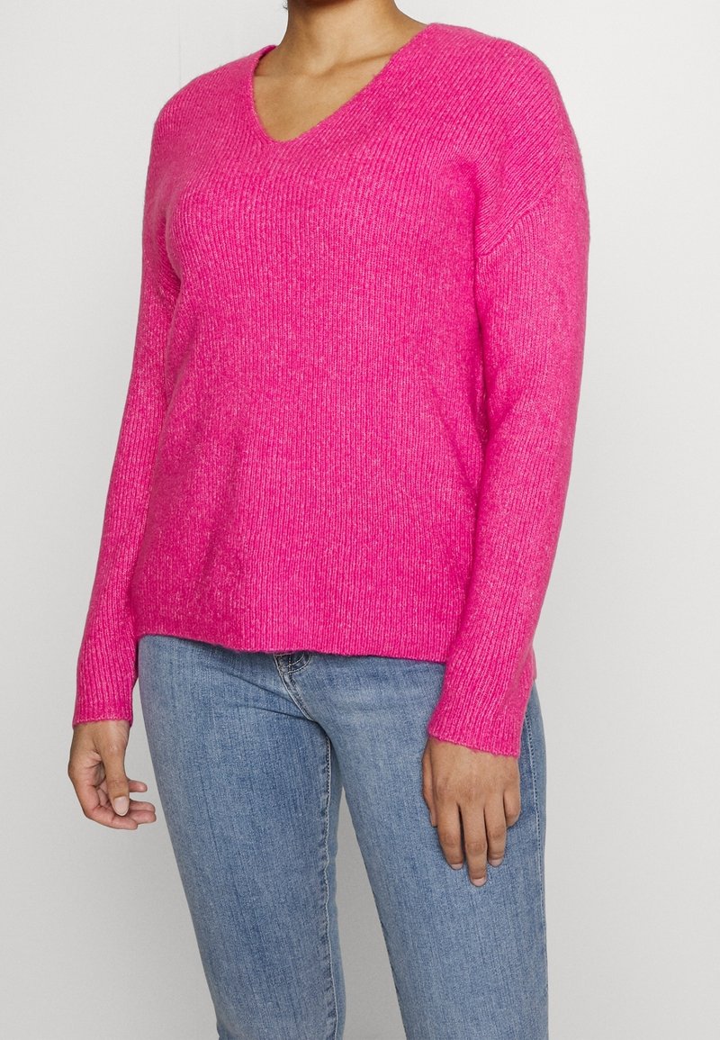 Rosa Pullover mit V-Ausschnitt, gerippter Textur, langen Ärmeln und einem leicht oversized Schnitt, kombiniert mit blauen Jeans.