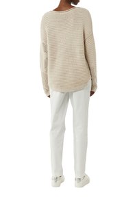 Beige Strickpullover mit lockerer Passform, der strukturierte, gewebte Ärmel und einen abgerundeten Saum hat, kombiniert mit weißen Hosen und weißen Sneakers.