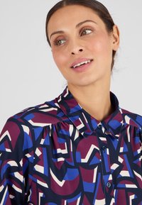 Blusa decorata con un mix di forme geometriche bordeaux, blu e crema. Design con colletto, chiusura a bottone e dettagli arricciati sulle spalle.