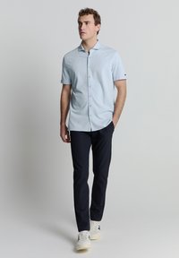 Helles blaues kurzärmliges Hemd mit Button-Down-Kragen, kombiniert mit dunkelblauen Hosen und weißen Turnschuhen. Glatte Textur, einfaches Design.
