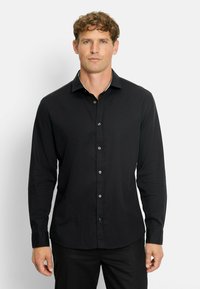 Mann mit lockigem Haar, der ein eng anliegendes schwarzes Langarmhemd und schwarze Hose trägt, steht vor einem weißen Hintergrund.