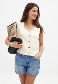 Beige stickad väst med fyra gyllene knappar och två framfickor, kombinerad med ljusblå denimshorts. Håller en strukturerad strapats och svart clutch.