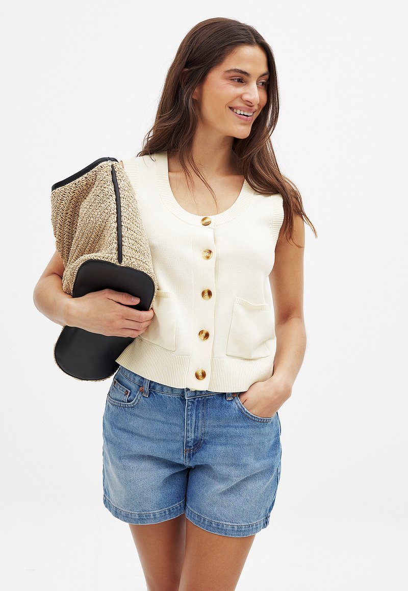 Beige stickad väst med fyra gyllene knappar och två framfickor, kombinerad med ljusblå denimshorts. Håller en strukturerad strapats och svart clutch.