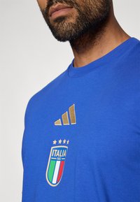 Μπλε βαμβακερό T-shirt με στρογγυλή λαιμόκοψη, που διαθέτει χρυσό λογότυπο της Adidas και κεντημένη ετικέτα της εθνικής ομάδας της Ιταλίας με αστέρια.
