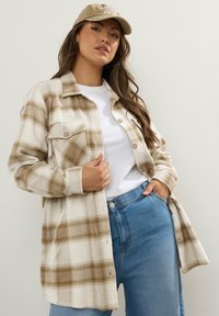 Beige ja cream ruuduline särk, millel on krae ja nööbiga kinnitamine, rinnatasku ja pehme tekstuur, sobitatud heleda sinise teksapüksi ja mütsiga.