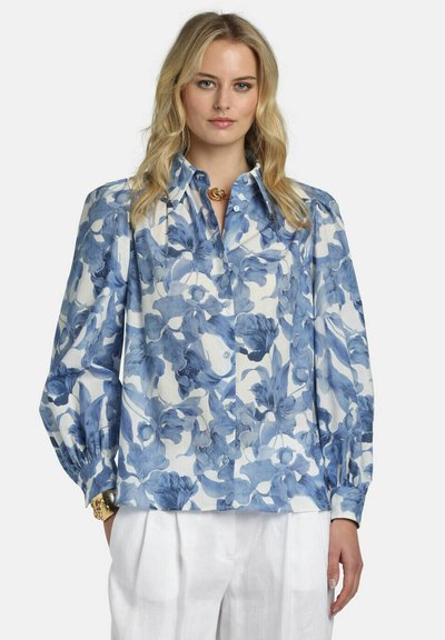 Woman wearing a blue and white floral button-up blouse with puffed long sleeves and white pants, accessorized with gold jewelry.