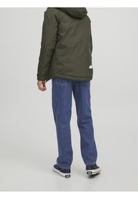 Persona che indossa una giacca con cappuccio verde scuro, jeans blu e sneakers nere, in piedi con la schiena rivolta verso la fotocamera su uno sfondo semplice.