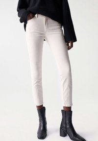 Cream-colored jeans with a fitted design, tapered leg, and subtle tonal stitching. Paired with black ankle boots featuring a block heel.