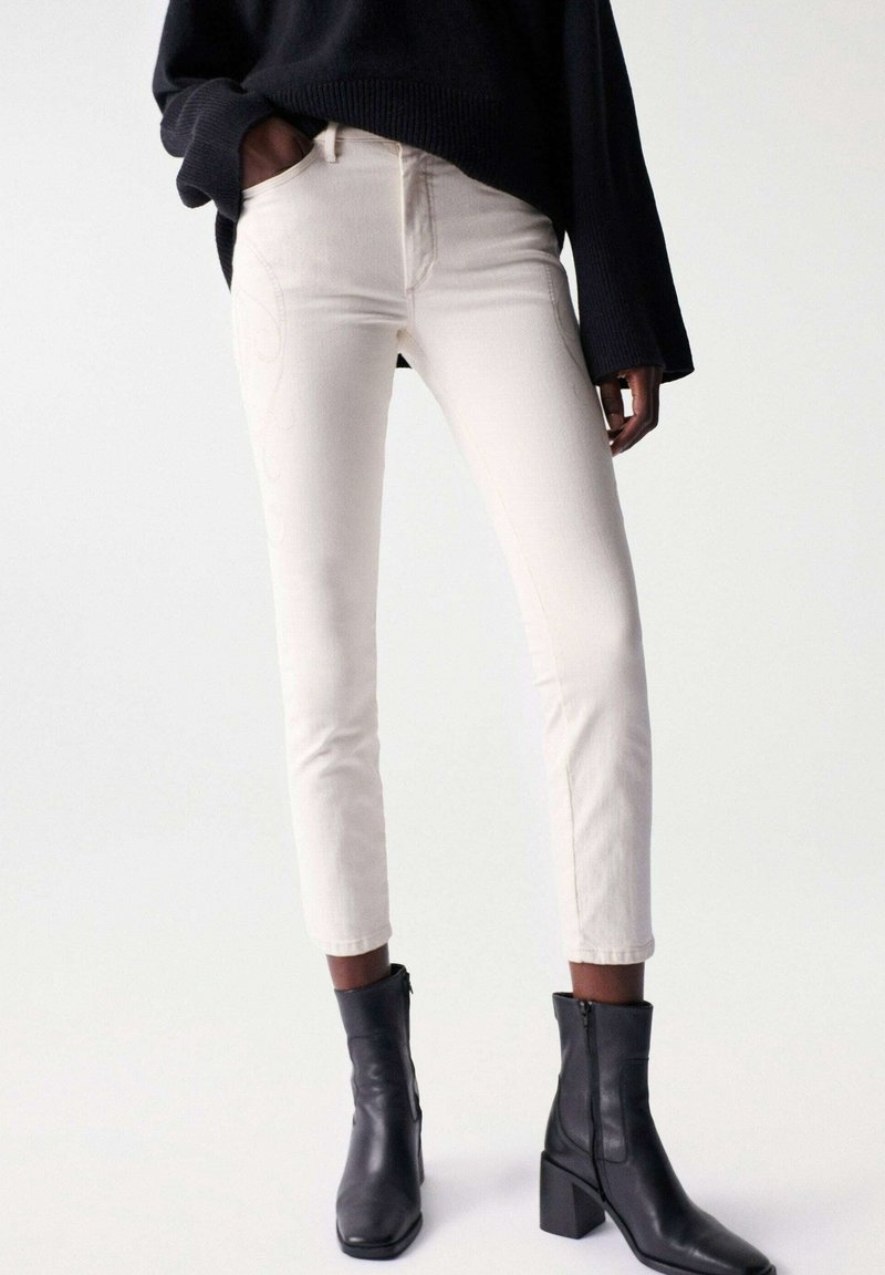 Cream-colored jeans with a fitted design, tapered leg, and subtle tonal stitching. Paired with black ankle boots featuring a block heel.