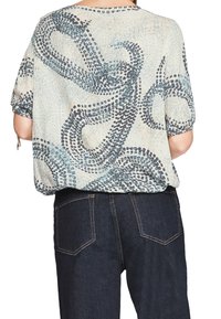 Blouse légère avec un motif abstrait bleu et gris, offrant une coupe ample et des manches courtes. Associée à un jean en denim foncé.