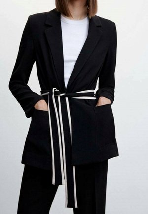 Kobieta ubrana w czarną marynarkę z białym paskiem z podwójnymi paskami zawiązanym w talii, w zestawie z białą koszulą i czarnymi spodniami. Ręce w kieszeniach.