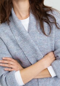 Cardigan lavorato a maglia blu chiaro con colletto a scialle, trama testurizzata e vestibilità rilassata. Indossato sopra una camicia bianca a maniche lunghe.