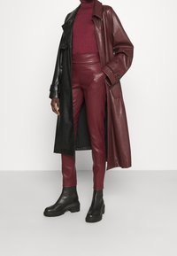 Abrigo largo de cuero negro sobre un cuello alto burdeos y pantalones de cuero burdeos ajustados. Botas de plataforma negras hasta el tobillo completan el look.