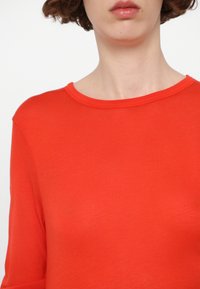 Červené triko s dlouhým rukávem a kulatým výstřihem, vyrobené z měkkého materiálu. Má hladký povrch a přiléhavý střih, ideální pro neformální nošení.