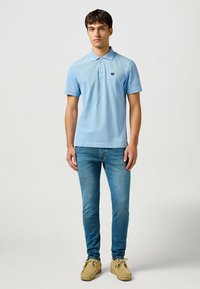 Ljusblå pikétröja med korta ärmar, en klassisk krage och en liten logotyp. Kombinerad med åtsittande blå jeans och beige skor.