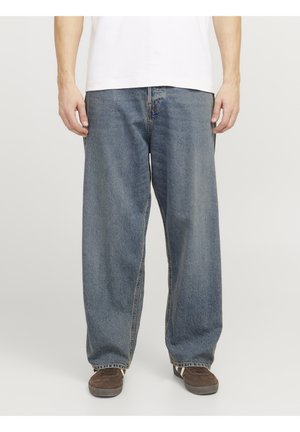 Personne portant un jean bleu large, une chemise blanche et des baskets marron, debout devant un fond uni.